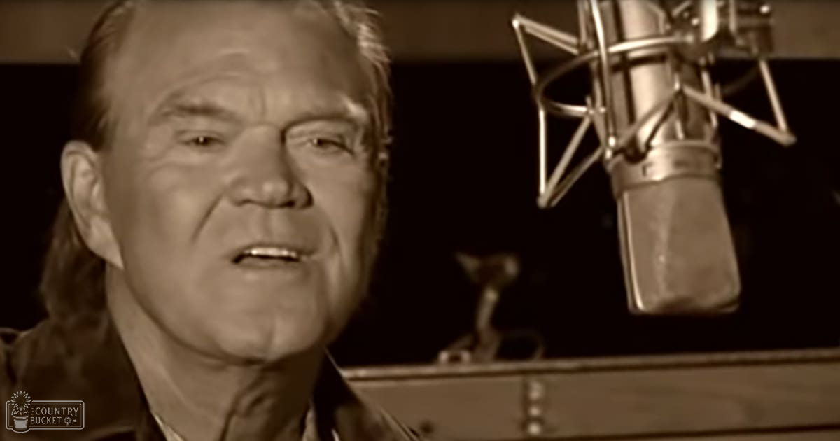 A middle-aged adult male wearing a dark-colored collared shirt with long sleeves and modern casual attire stands in a recording studio setup.