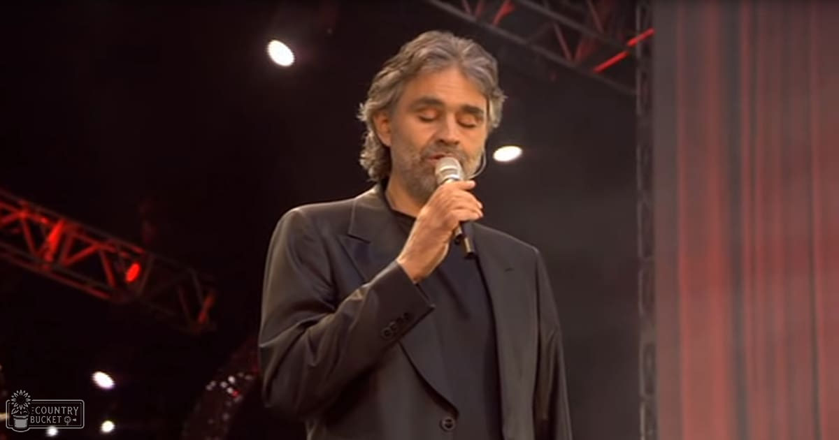 Middle-aged male singer performing on a dark stage with professional lighting, wearing a double-breasted suit and black shirt. Red stage lights highlight his presence in an indoor performance venue.
