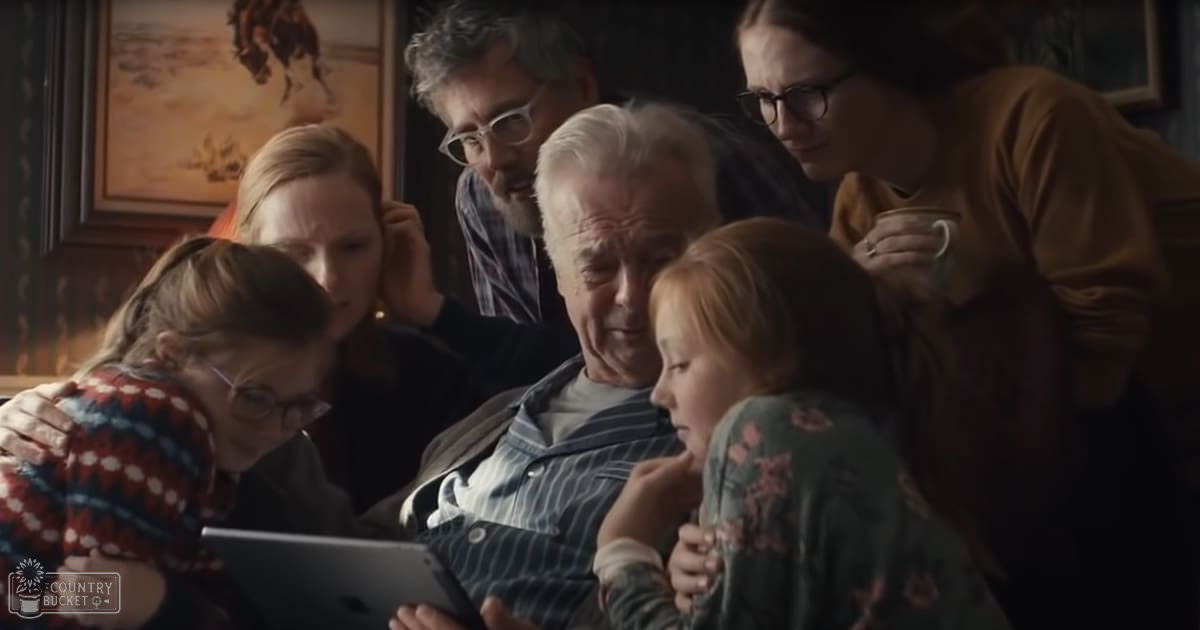 A family of four gathers around an older adult holding a tablet device in a dimly lit home interior with framed artwork on dark walls.