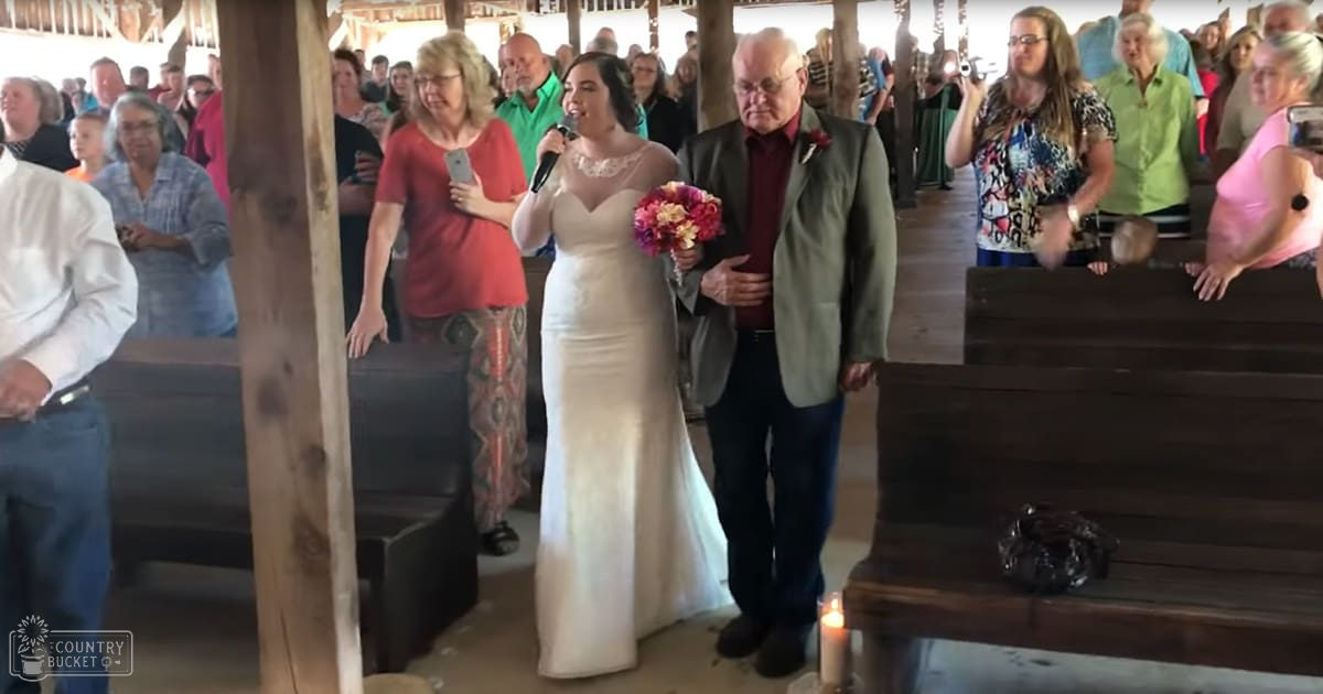 A young adult female bride in a white wedding gown walks down an aisle with her older male father wearing a dark green suit, under soft daylight at what appears to be a small church or similar venue.