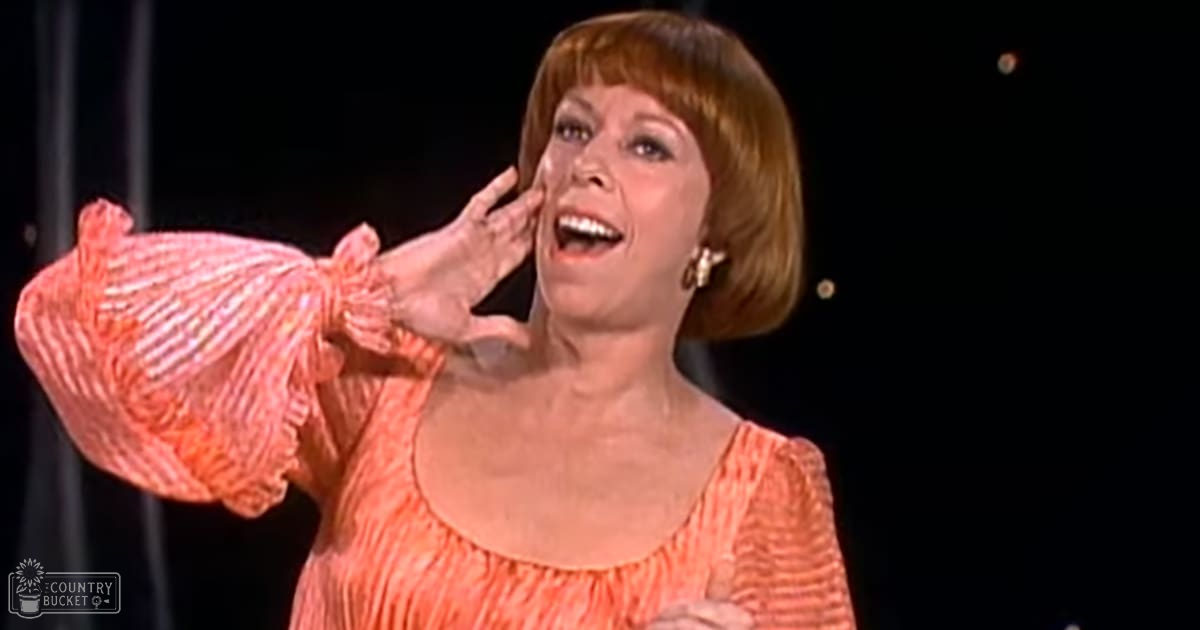 A middle-aged adult female wearing a coral-pink, striped, ruffled-sleeved dress with gold earrings, performing indoors under low-light conditions.