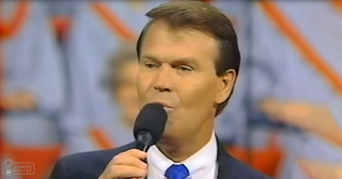 A middle-aged man speaks into a microphone against an indoor backdrop with orange and blue tones, suggesting a formal setting such as a conference or television studio.