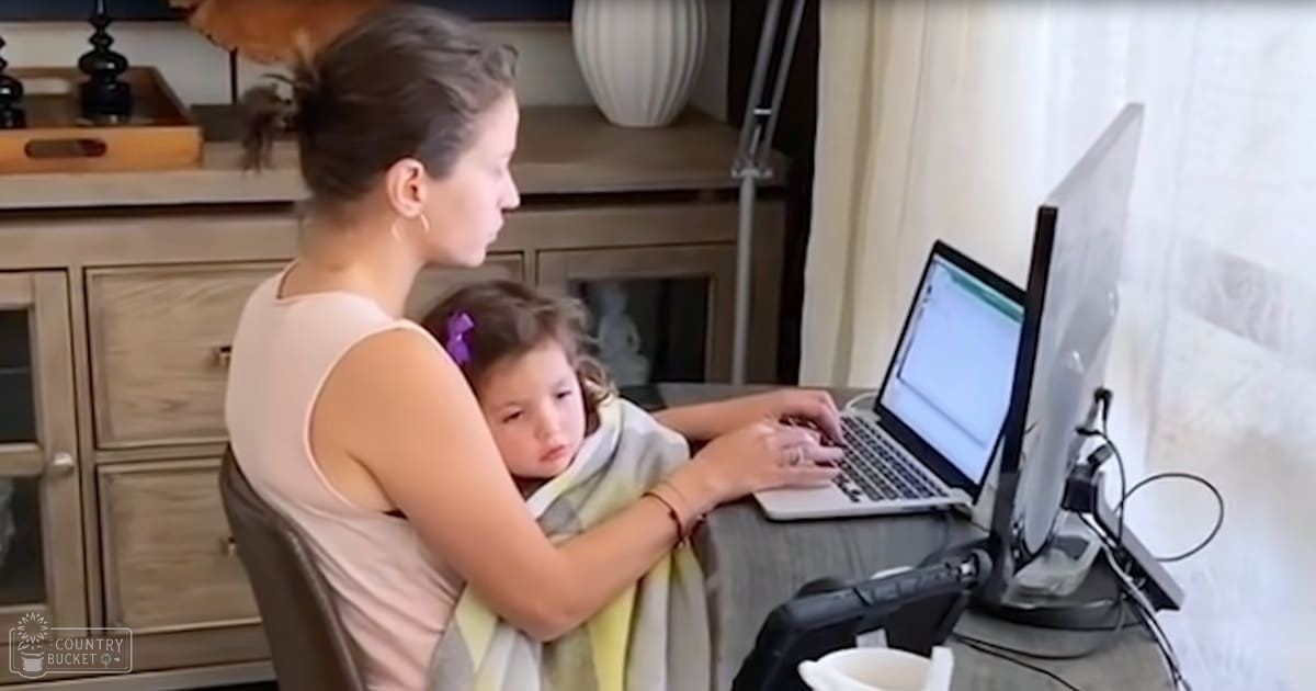 A young adult female wearing a light pink sleeveless top with earrings and bracelet, holding a toddler girl in her lap. The scene is set in a home office or study area with natural daylight.