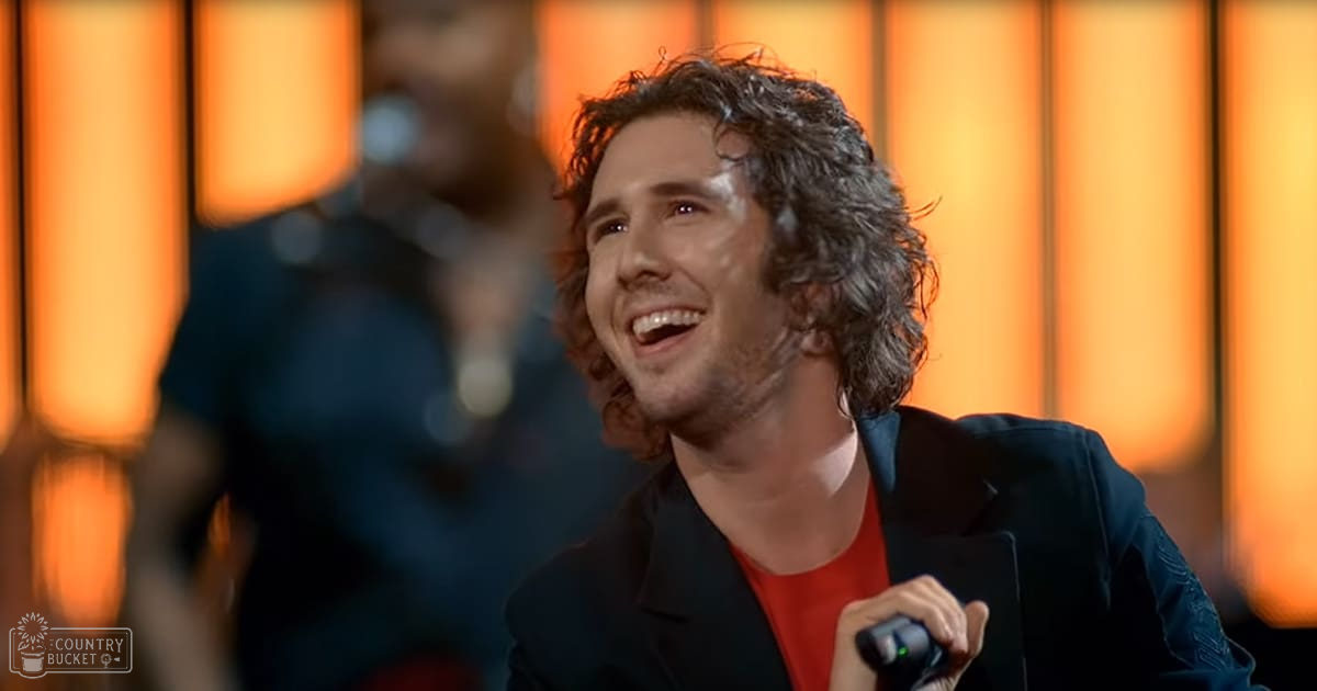A young adult male singer performing on stage wearing a dark blazer over a red t-shirt with visible embroidery, holding a microphone.