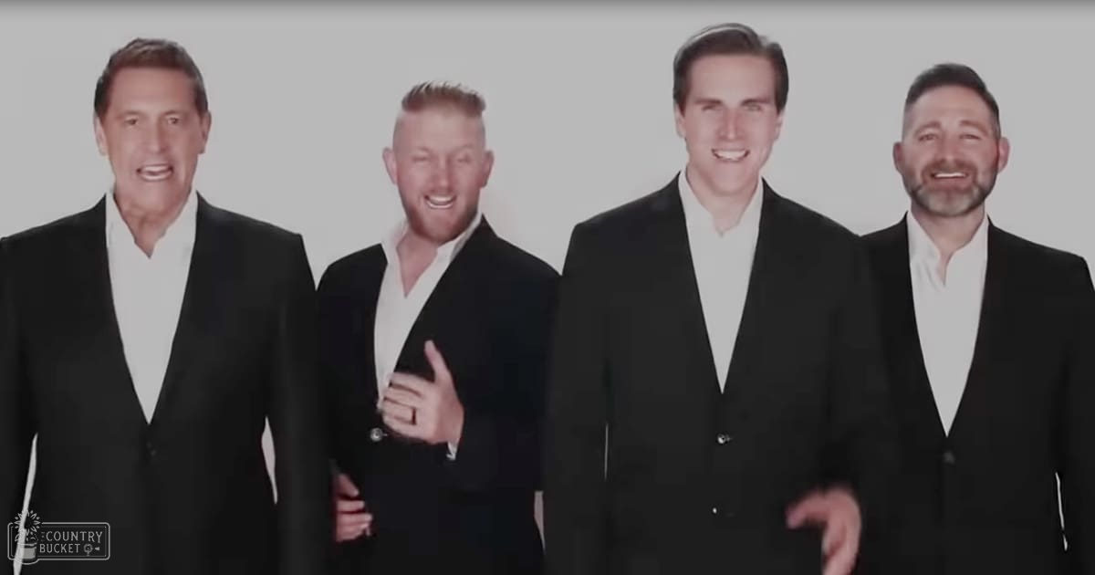 Four adults in formal attire standing indoors, wearing tailored black suit jackets over white collared shirts under soft indoor lighting.