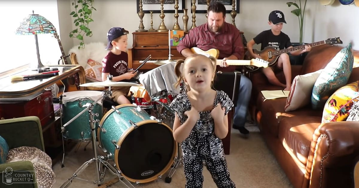 A young girl wearing a black and white patterned jumpsuit stands in front of a boy playing drums. An adult male plays guitar while seated at a piano bench, with decorative items like a stained glass lampshade and leather couch visible in the background.