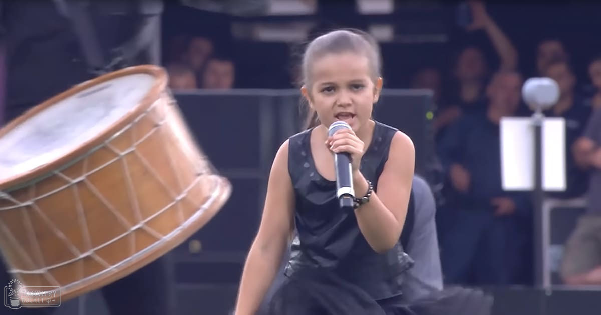 A young girl, approximately 8-10 years old, wearing a sleeveless black dress with ruffled details and standing on stage at an outdoor concert or festival.