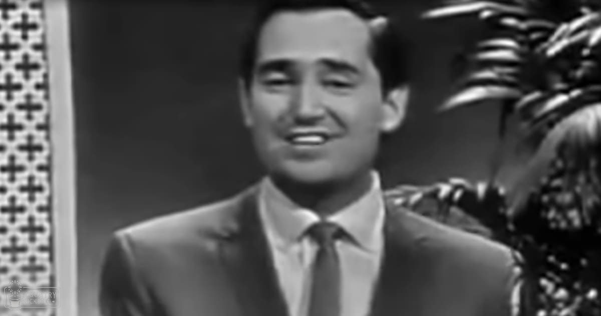 A man in a well-fitted suit jacket with notched lapel, white dress shirt, and dark tie stands indoors against patterned wall.