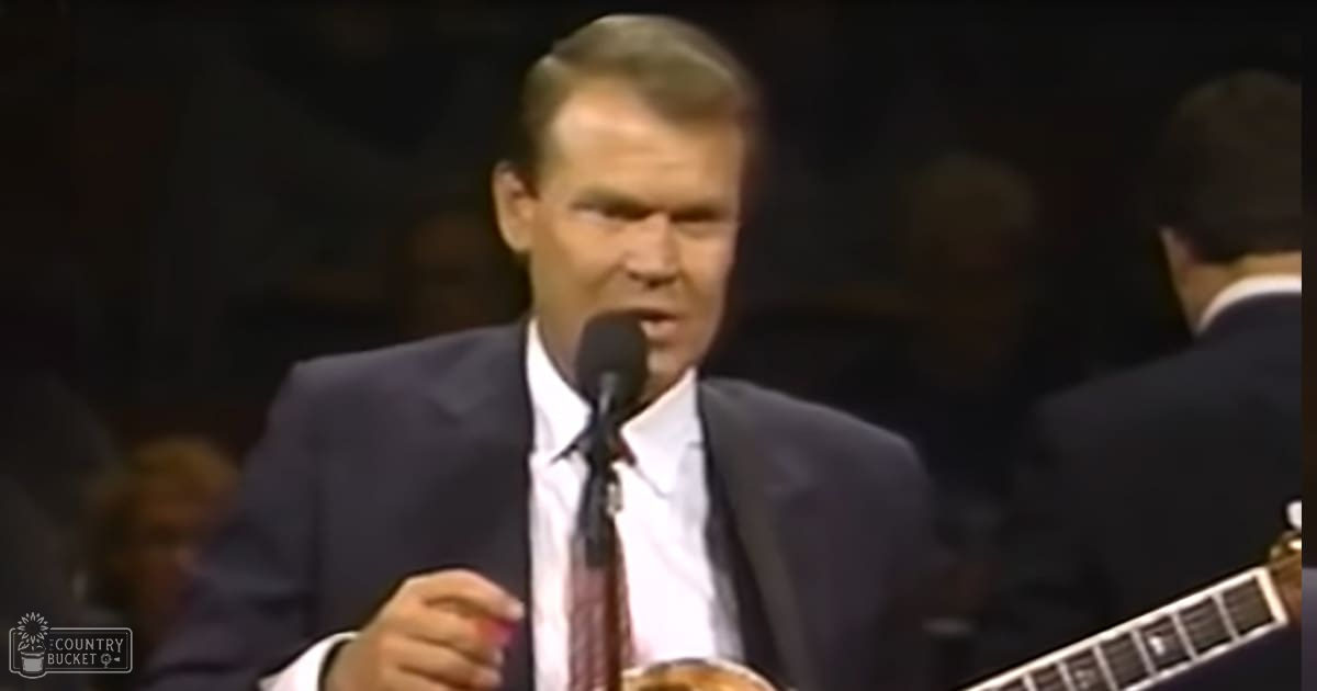 A middle-aged man in formal attire holds a microphone indoors, suggesting a performance or event setting.