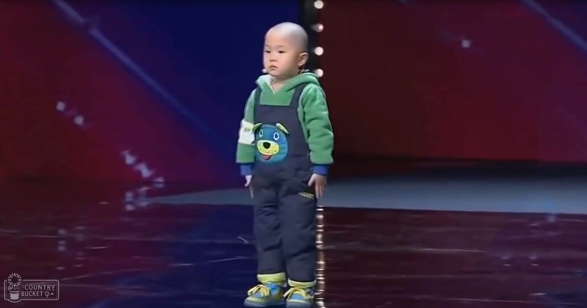 A toddler in a casual outfit stands on stage under red lighting, suggesting an indoor performance venue.