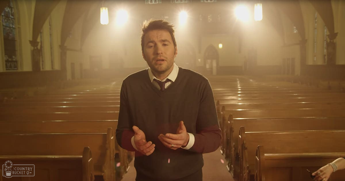 An adult male in a church setting, wearing a dark brown sweater over a white collared shirt with rolled-up sleeves and a maroon tie. The wooden pews line the aisle, while stained glass windows are partially visible on the left side.