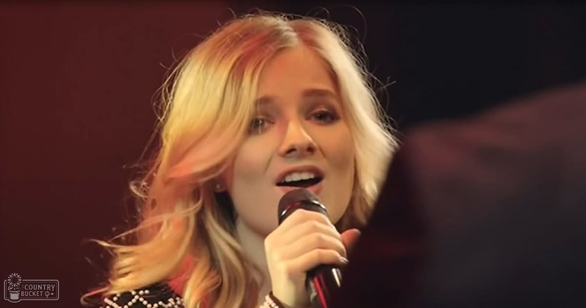 A young adult female singer performing at an indoor event, wearing a dark patterned top and holding a microphone against a dimly lit background suggesting evening or night time.
