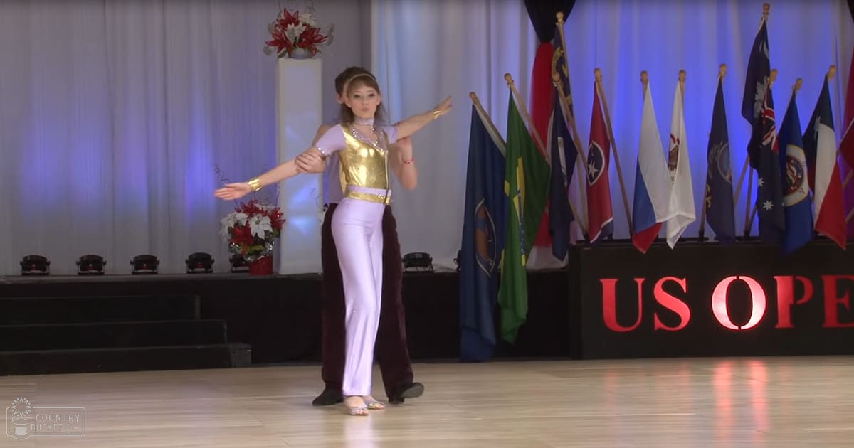 A young adult female dancer wearing a gold leotard and white pants, performing at an 'US Open' dance competition with flags from various countries in the background.