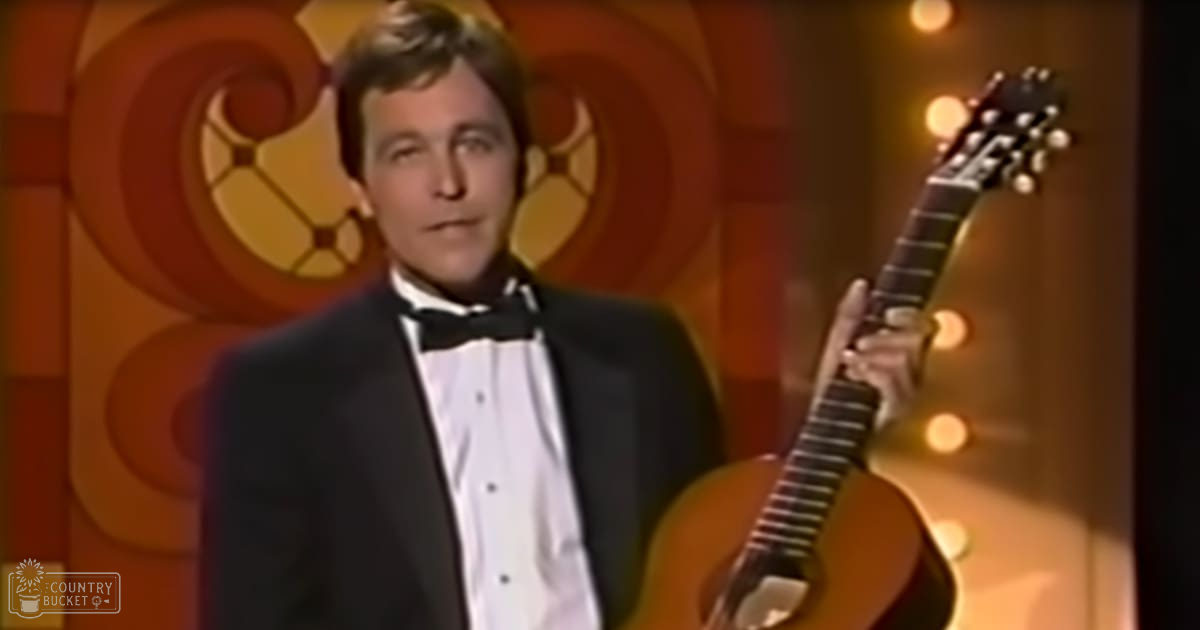 An adult male singer in a formal tuxedo with a bow tie, holding a guitar on an indoor stage with warm-toned decorative walls.