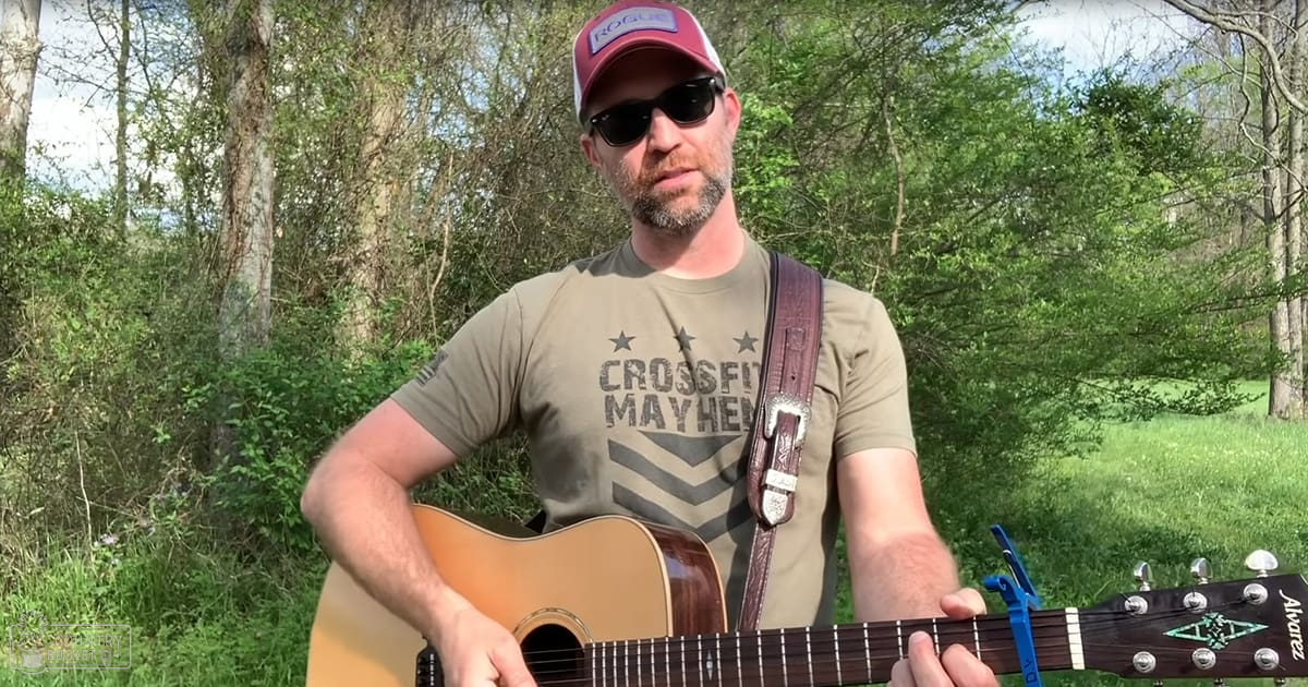 Adult male playing acoustic guitar in a wooded outdoor setting with green grass and leafy trees, suggesting spring or summer day.