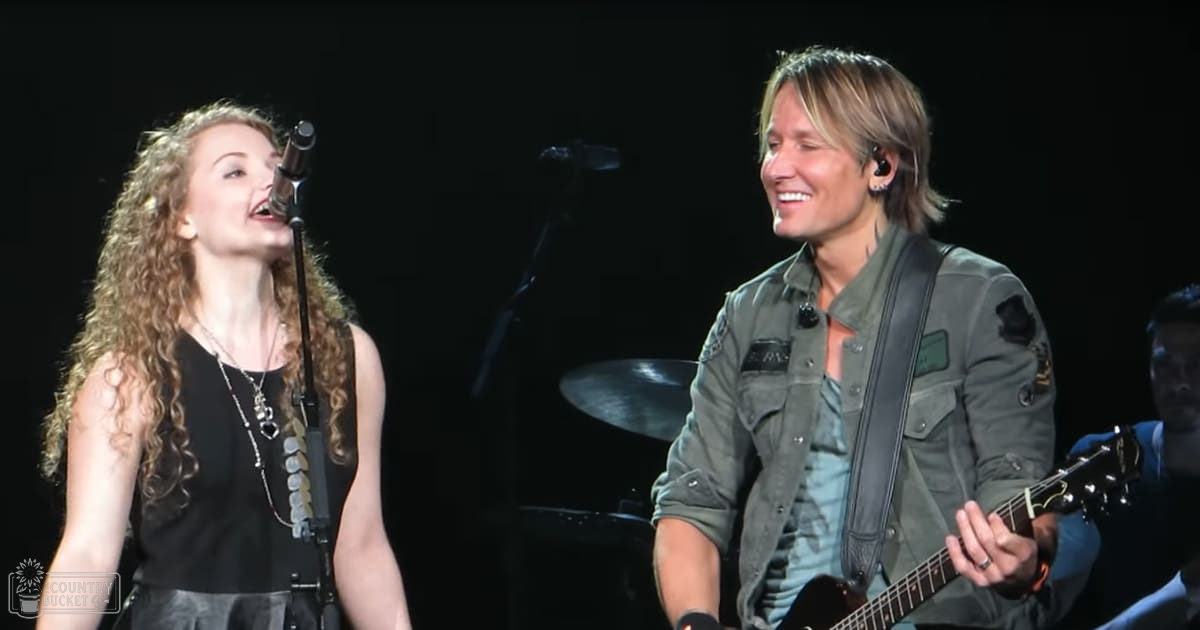 Young adult female singer wearing a black sleeveless top and layered necklaces, performing on stage at an indoor concert venue alongside an adult male guitarist in an olive green jacket with patches.