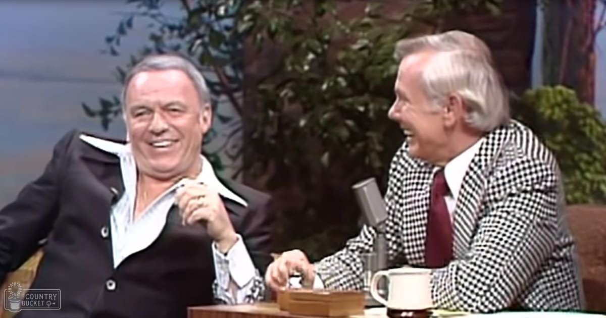 Middle-aged man with short gray hair wearing a black blazer over a white shirt with an open collar and dark tie, interviewed by another older adult in a checkered jacket and maroon tie in a talk show studio setting.