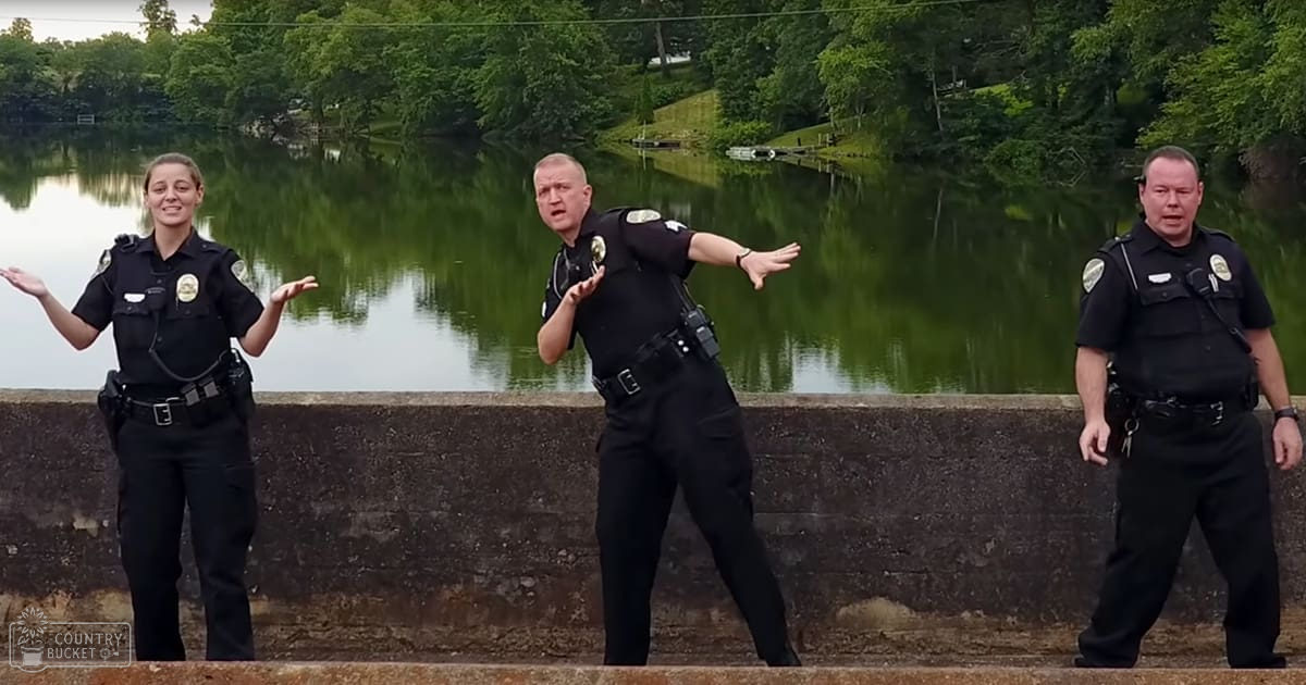 A group of police officers in uniform standing by a lake at what appears to be an outdoor park during spring or summer, with lush greenery and soft daylight.