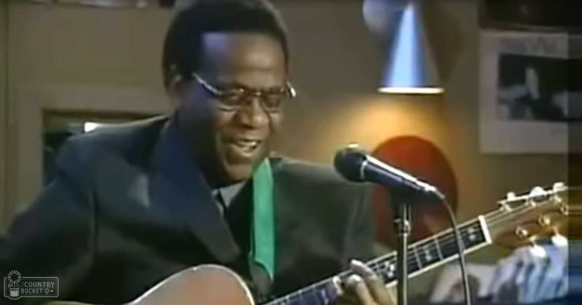 An adult male musician performing in a recording studio, wearing a dark suit jacket with a green scarf and glasses, surrounded by an acoustic guitar, microphone, lamp, and framed picture on the wall.