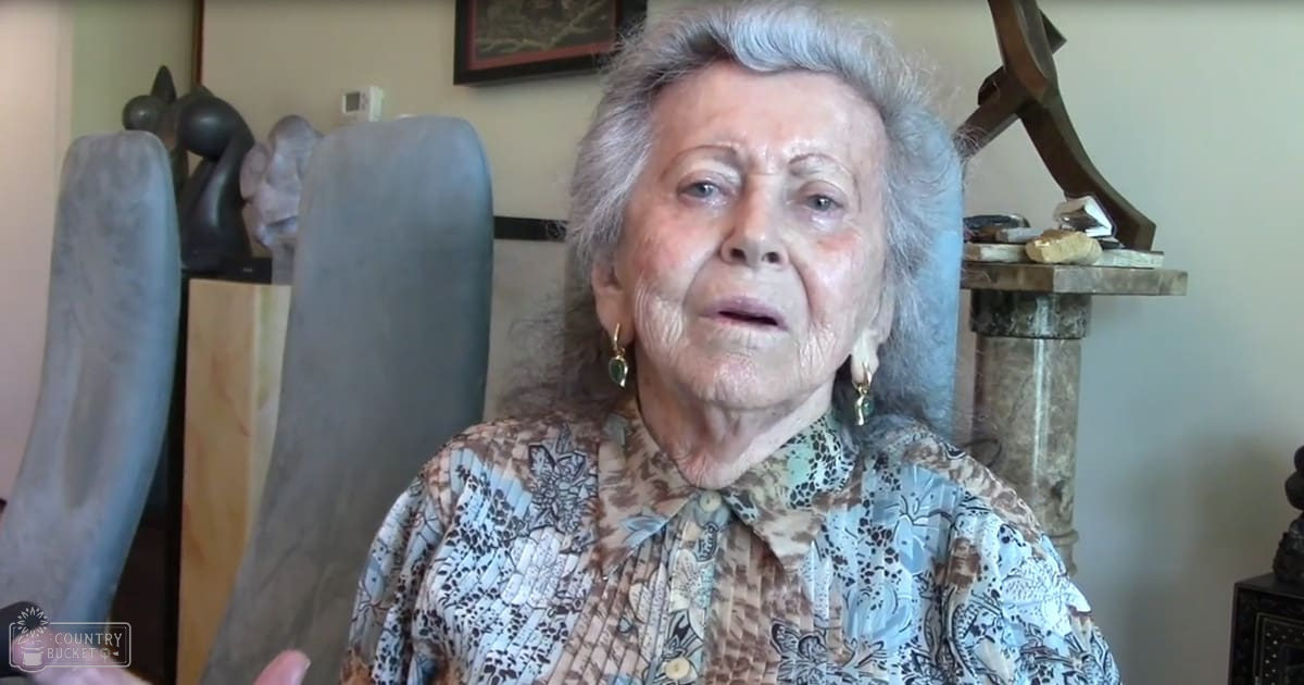 An older adult female in a home setting, wearing a patterned blouse with gold hoop earrings. A dark-colored chair and framed picture are visible in the background.