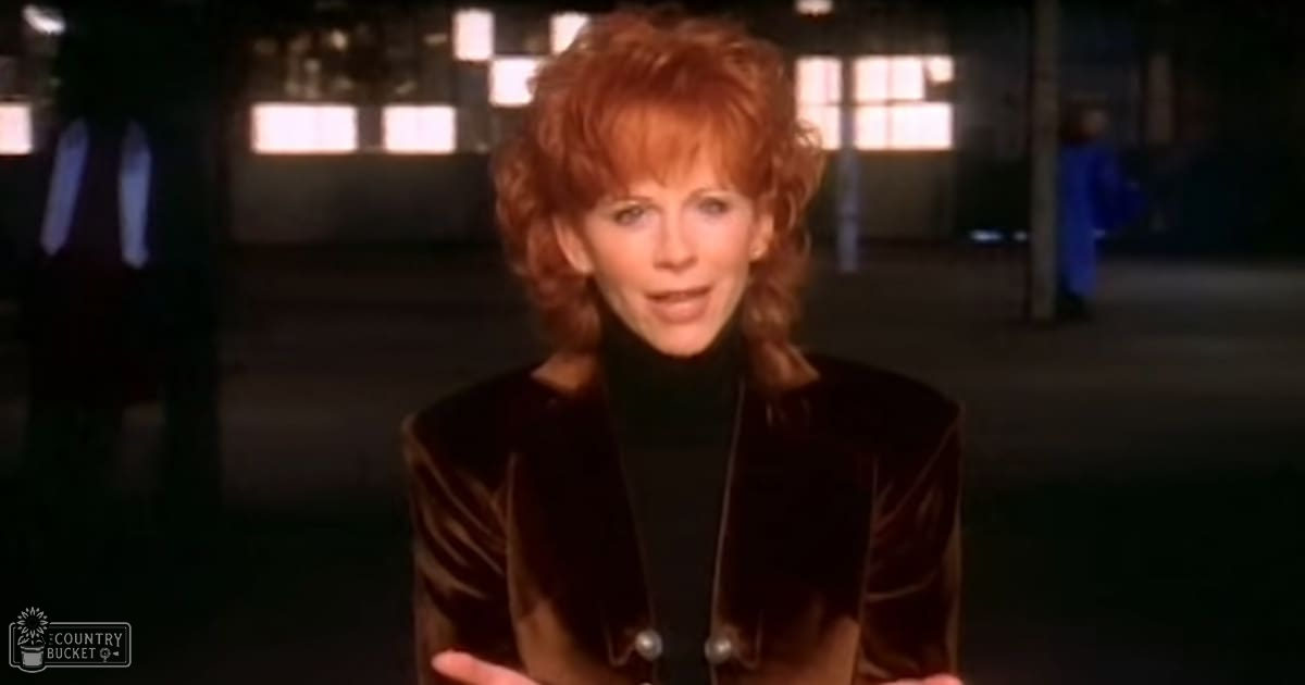 A middle-aged woman in a relaxed fit, dark-colored jacket with high collar and button closure at an industrial or warehouse-like setting.