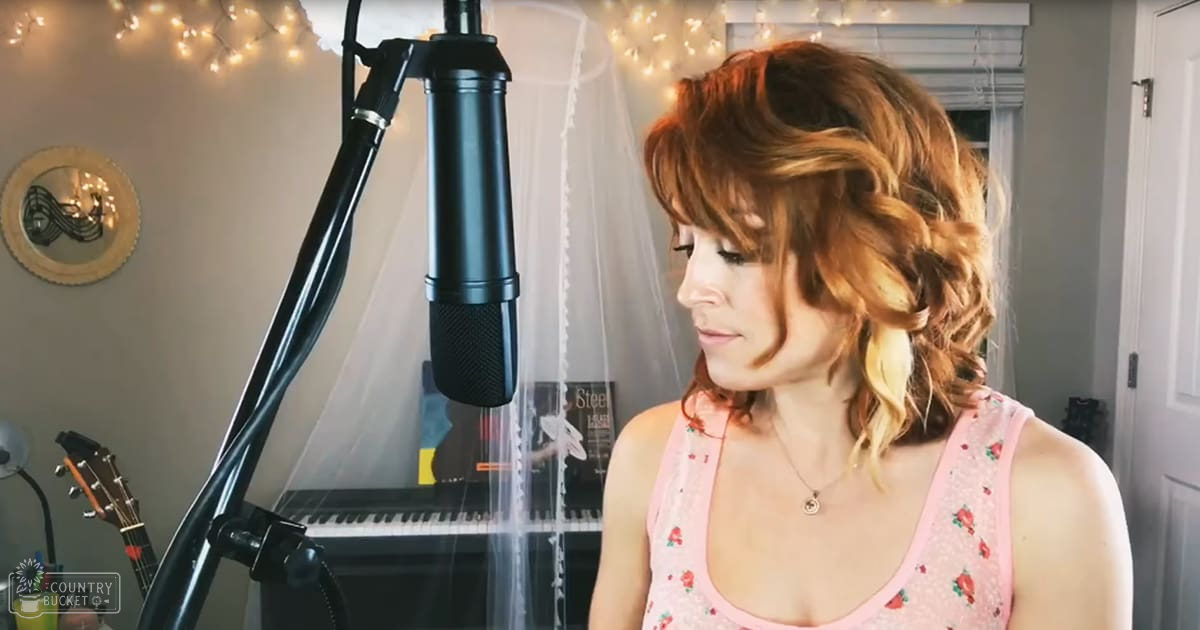 A young adult female wearing a pink sleeveless top with floral pattern and lace detailing, standing in a home recording studio setting.