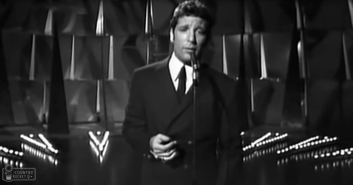A man in an adult age band wearing a dark suit jacket with a white shirt and tie stands behind a microphone on a stage, suggesting he is performing or speaking at an event.
