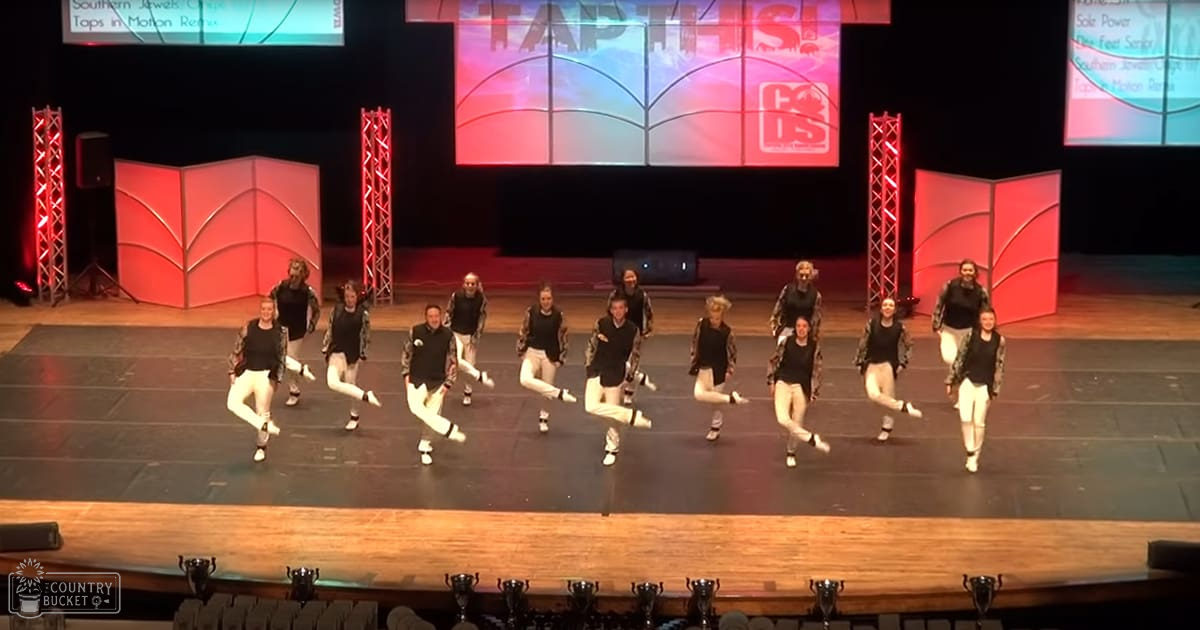 A dance group of young adult females performing on stage at an event with a red-lit backdrop in an indoor auditorium or theater setting.