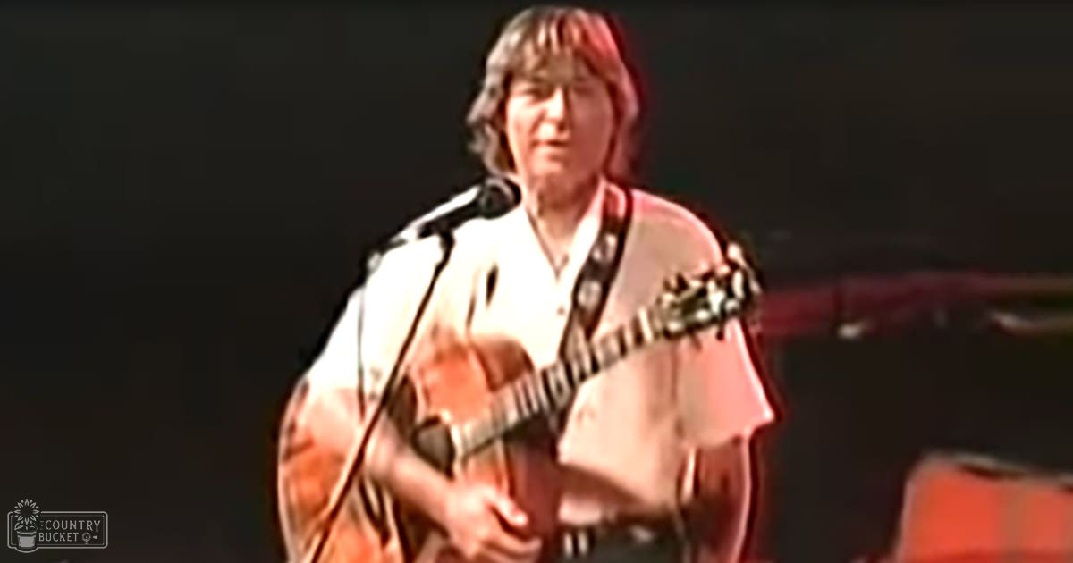 A middle-aged male musician in a white shirt performs on stage with an acoustic guitar, under focused lighting against a dark backdrop.