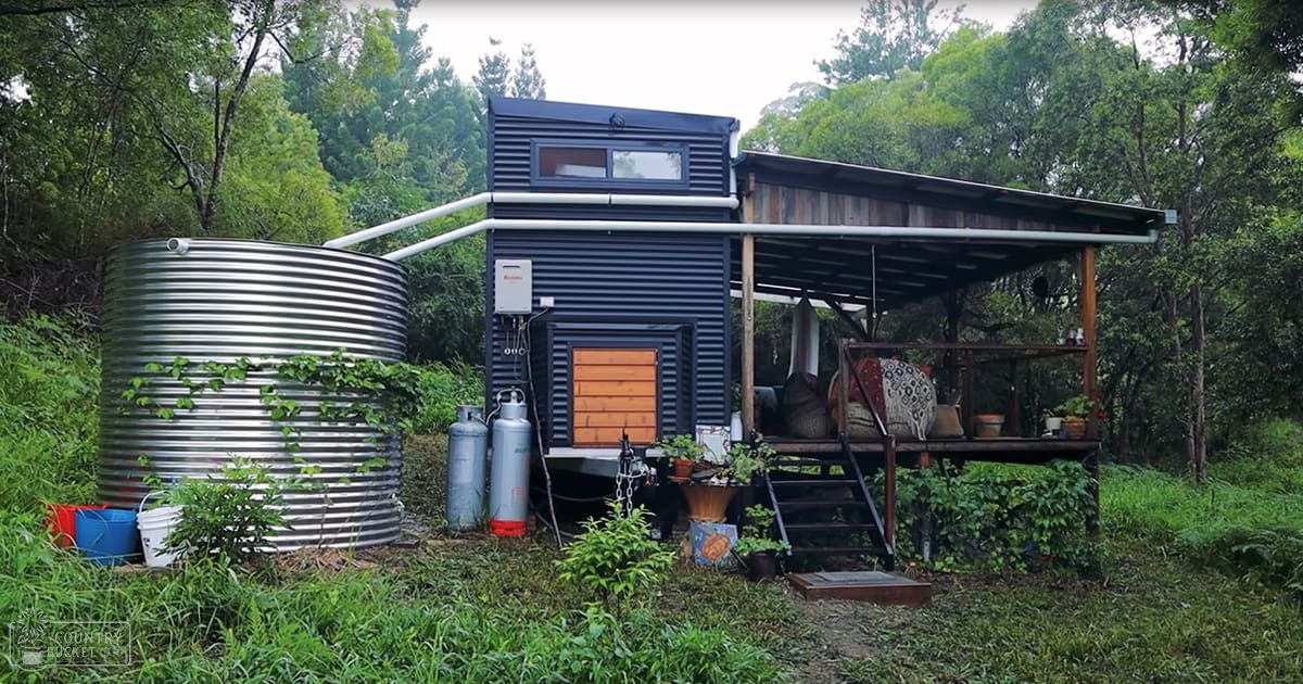 A serene forested area with a small modern house, water tank, propane tanks, outdoor seating, and lush greenery in spring or summer light.