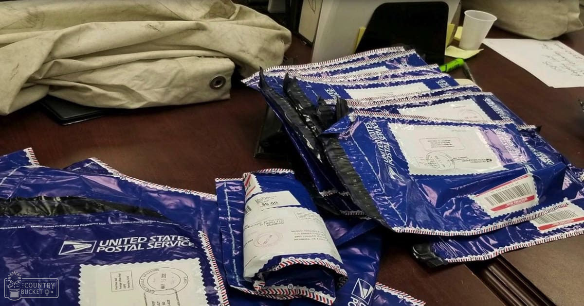 Stacked United States Postal Service packages on a dark wooden table in an indoor office environment with blue, white, beige, black, and brown colors dominating the scene.