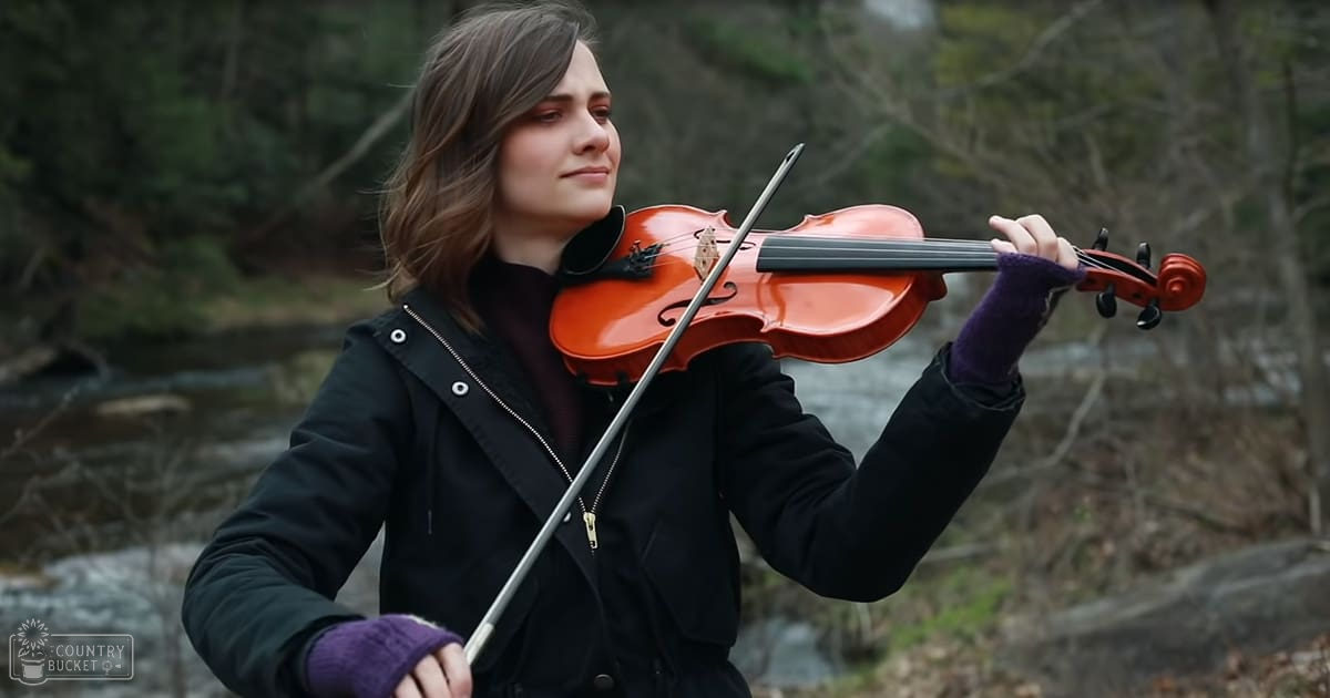 A young adult female playing the violin outdoors near a body of water, dressed in a dark-colored jacket with purple fingerless gloves.