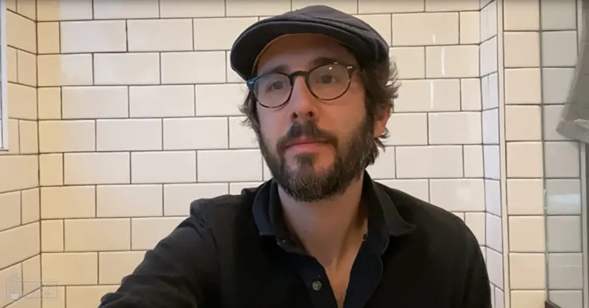 An adult male in a kitchen setting, wearing a dark-colored button-up shirt with round glasses and a flat cap.