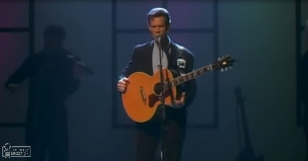 Adult male performer wearing dark suit and light shirt on stage with an acoustic guitar, geometric backdrop at night.