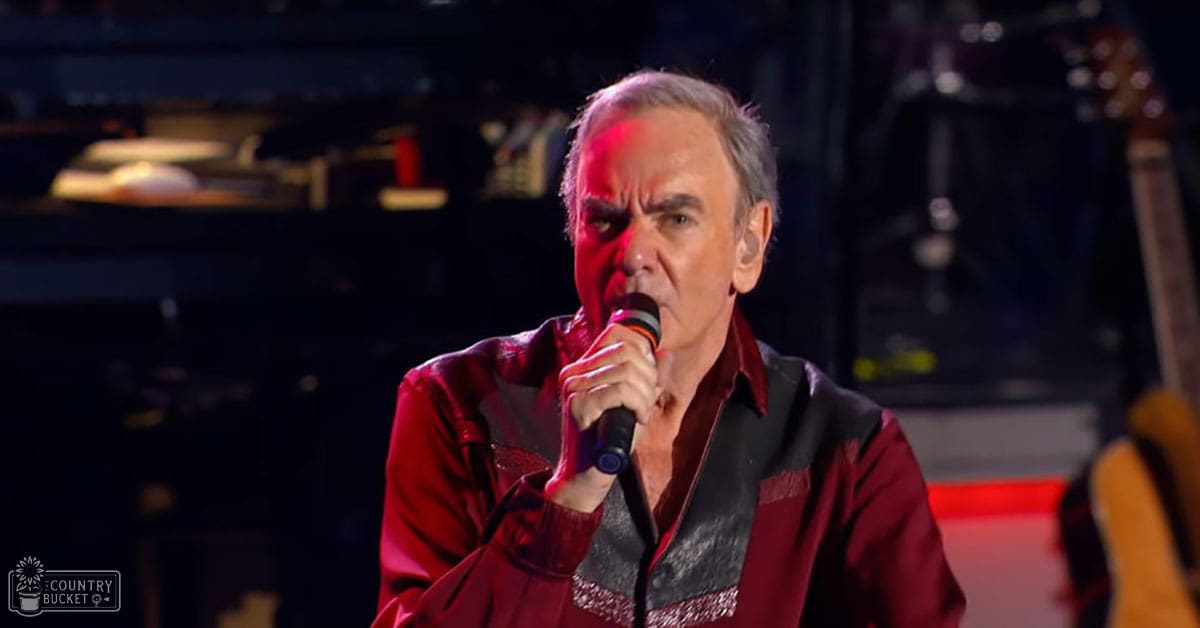 A middle-aged adult male in a red jacket with black accents performs on an indoor concert venue under dim stage lighting.