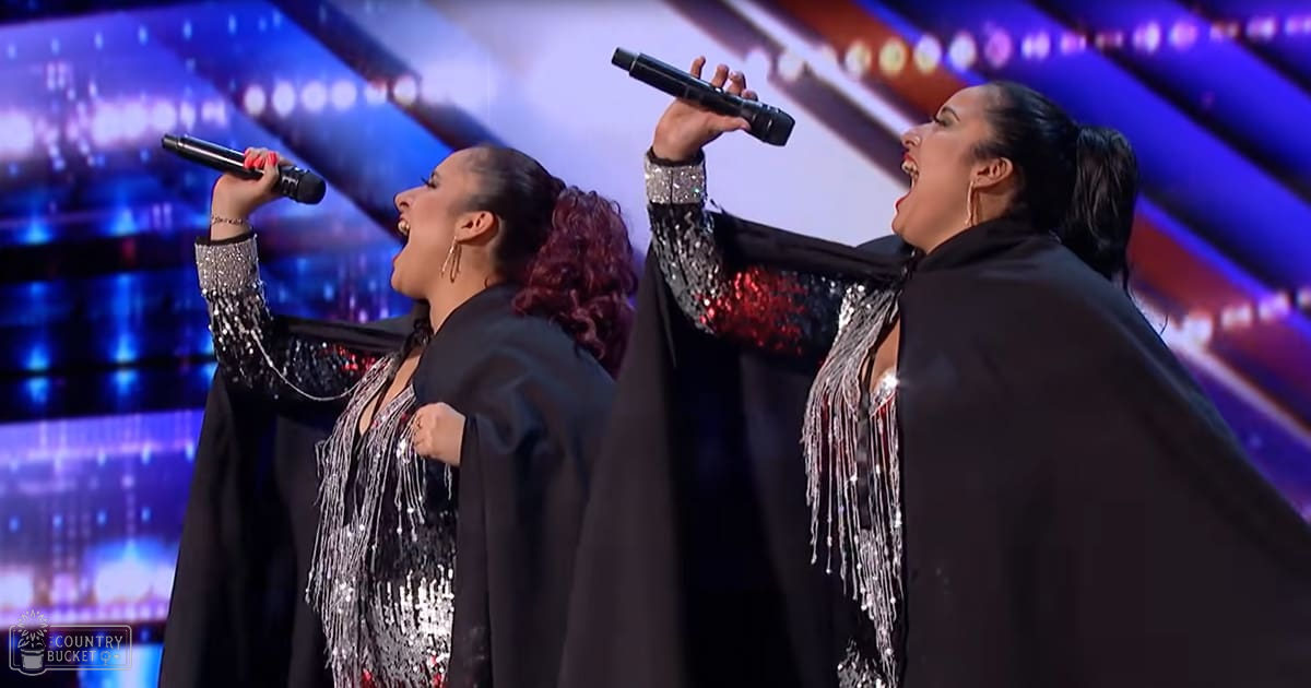 Two adult female performers singing on a stage with vibrant lighting at an indoor concert or talent show, wearing matching black sequined capes and dresses.