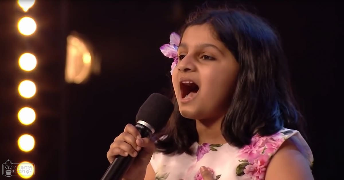 A young girl in a white floral dress sings into a microphone during what appears to be a stage performance, illuminated by stage lighting against a dark background.