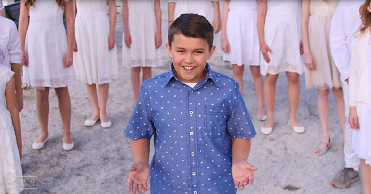A young boy wearing a blue short-sleeved button-up shirt with white polka dots stands outdoors on sandy ground surrounded by children dressed in white dresses, suggesting it's late afternoon during spring or summer.
