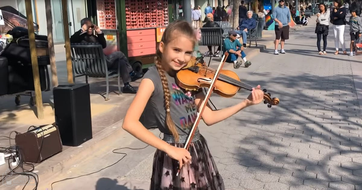 A young girl playing a violin outdoors in an urban pedestrian area with benches and people sitting nearby.