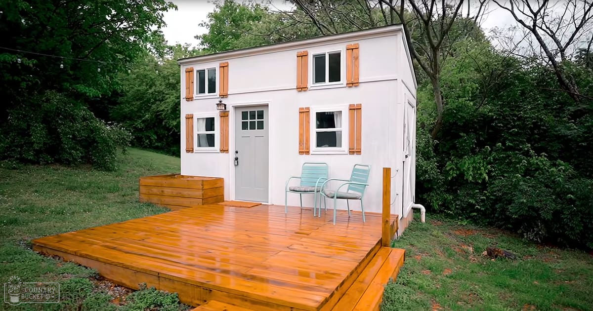 A small white cabin with wooden shutters surrounded by lush greenery in an outdoor residential area during spring or summer, under overcast skies.