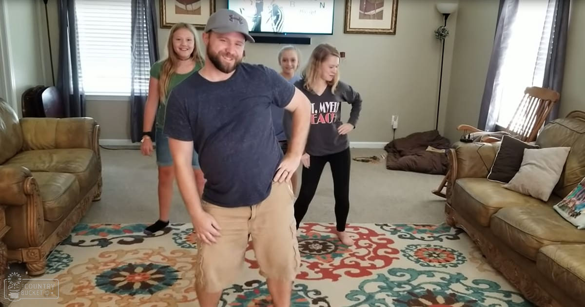 A middle-aged adult male in a living room with beige furniture, floral-patterned rug, and two younger females. The setting is daytime indoors.