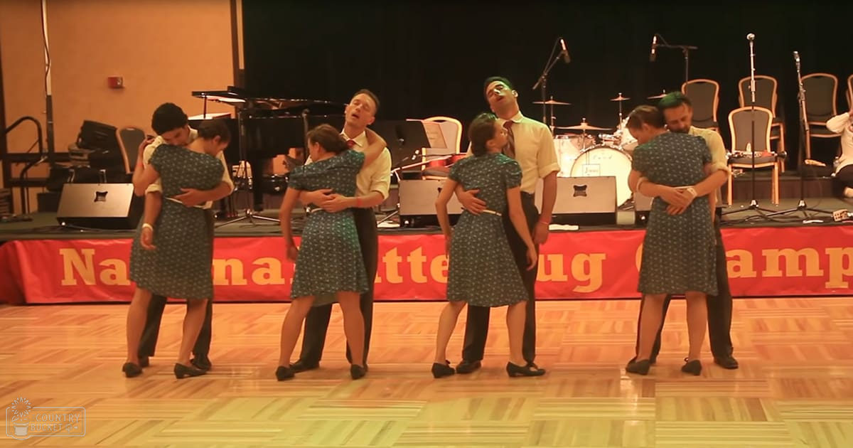A group dance performance with young adults in dark green dresses, set against a stage with musical instruments and warm-toned lighting.