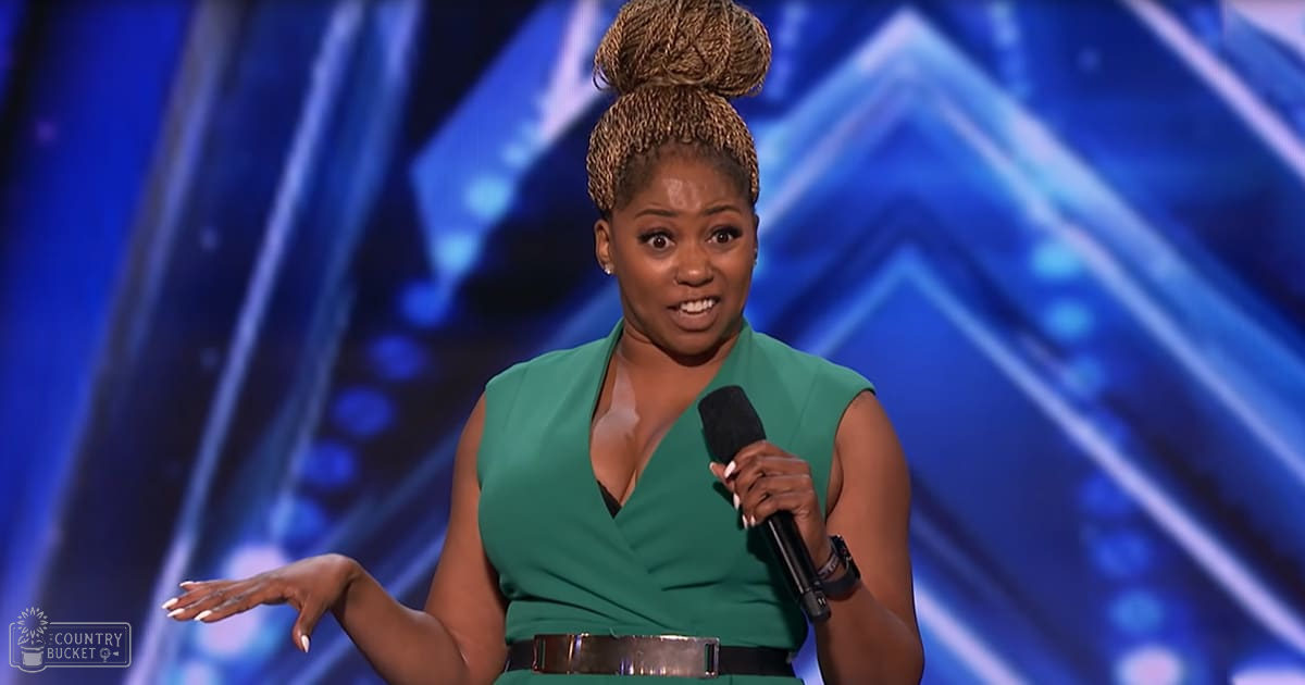 An adult female performer in a modern green sleeveless dress with a deep V-neckline, holding a microphone on an indoor stage with blue-toned geometric backdrop and stage lighting.