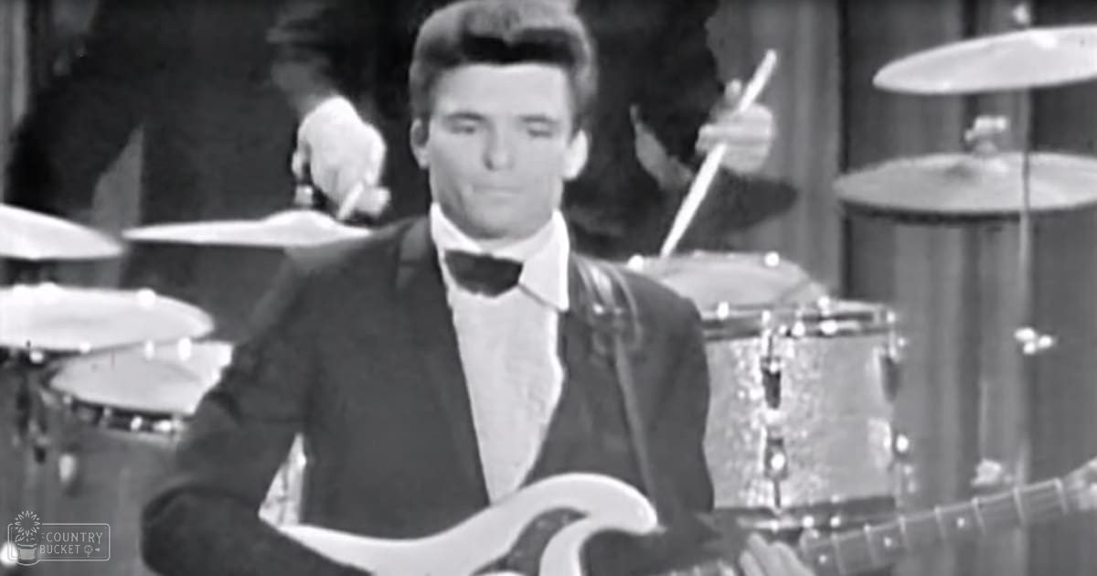 A young adult male wearing a dark suit jacket and white shirt with a bow tie plays guitar on stage in an indoor performance setting, illuminated by overhead stage lights.