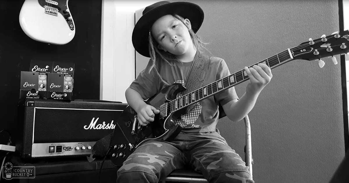 A child wearing a dark gray t-shirt with camouflage pants, playing guitar in a music studio or practice room with a Marshall DSL 100H amplifier and Elixir guitar strings visible.