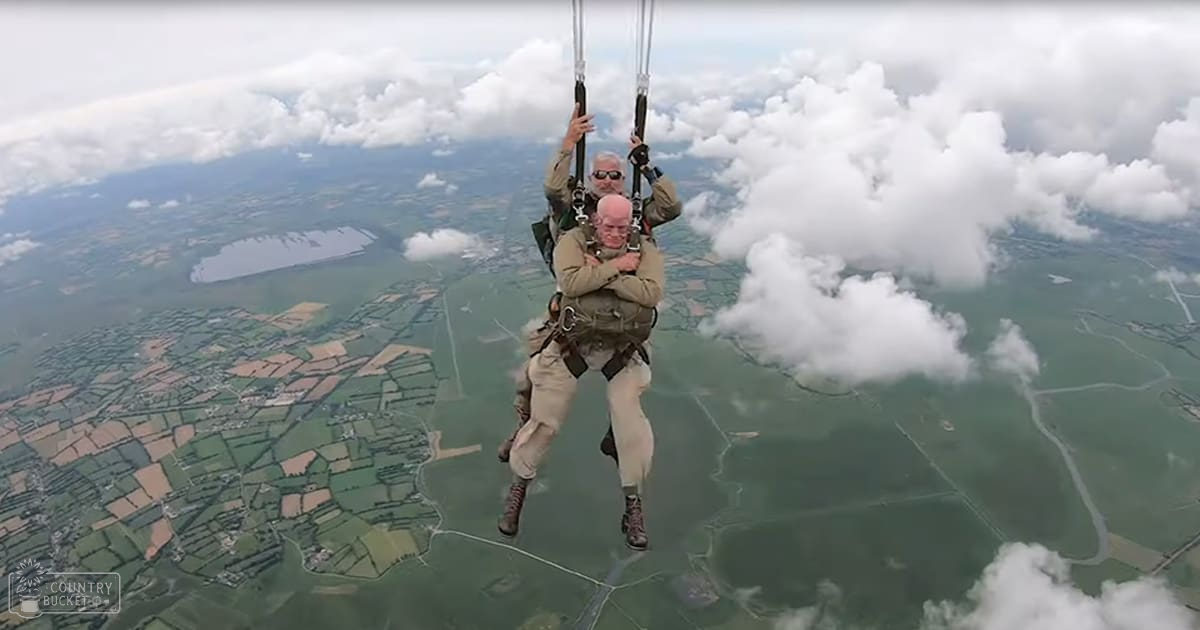 Two skydivers in mid-air over a landscape with fields and water bodies, surrounded by clouds. The season appears to be spring or summer due to the greenery and lack of snow.