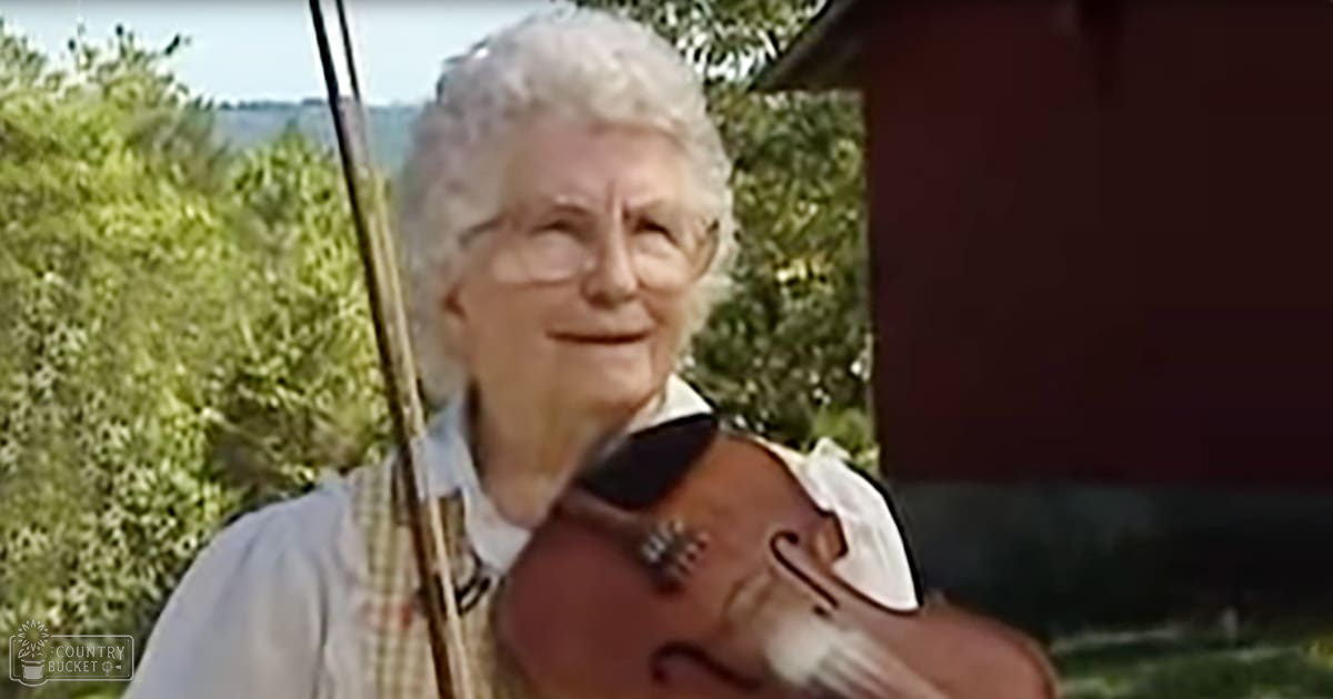 An older adult female holding a violin outdoors in a rural setting with greenery and trees, and a red structure nearby.