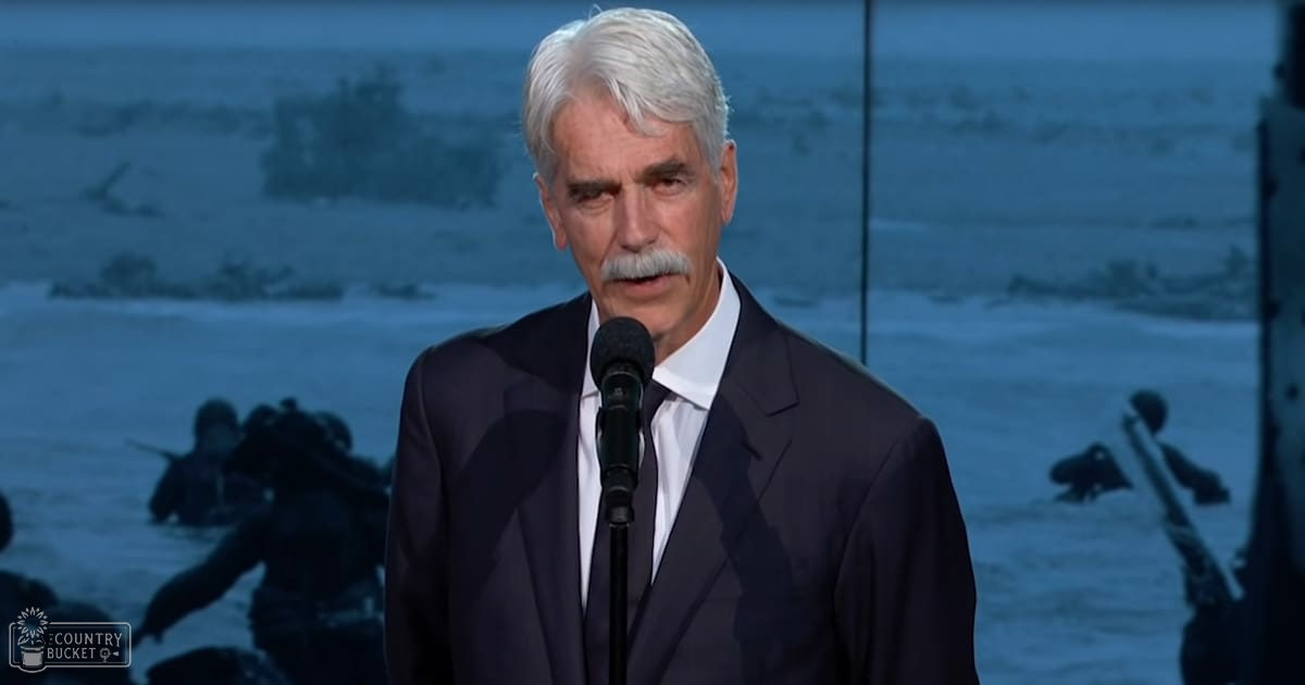 A middle-aged man in a formal dark suit speaking at an event with a somber military-themed backdrop, likely related to remembrance.