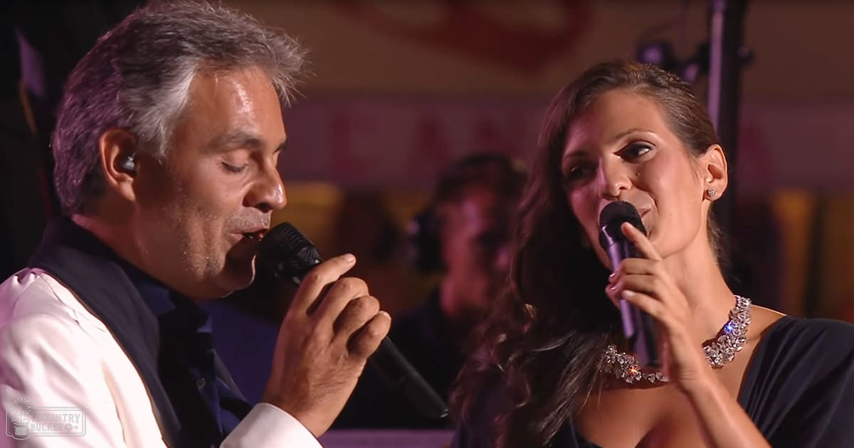 A musical performance scene at an indoor venue with a male adult with graying hair wearing a white shirt under a dark vest and a female adult with long dark hair in a black dress with a large stone or crystal necklace.