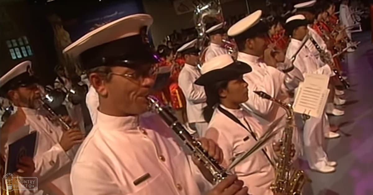 A marching band performance with musicians in white uniforms, playing various brass and percussion instruments under stage lighting in an indoor venue.